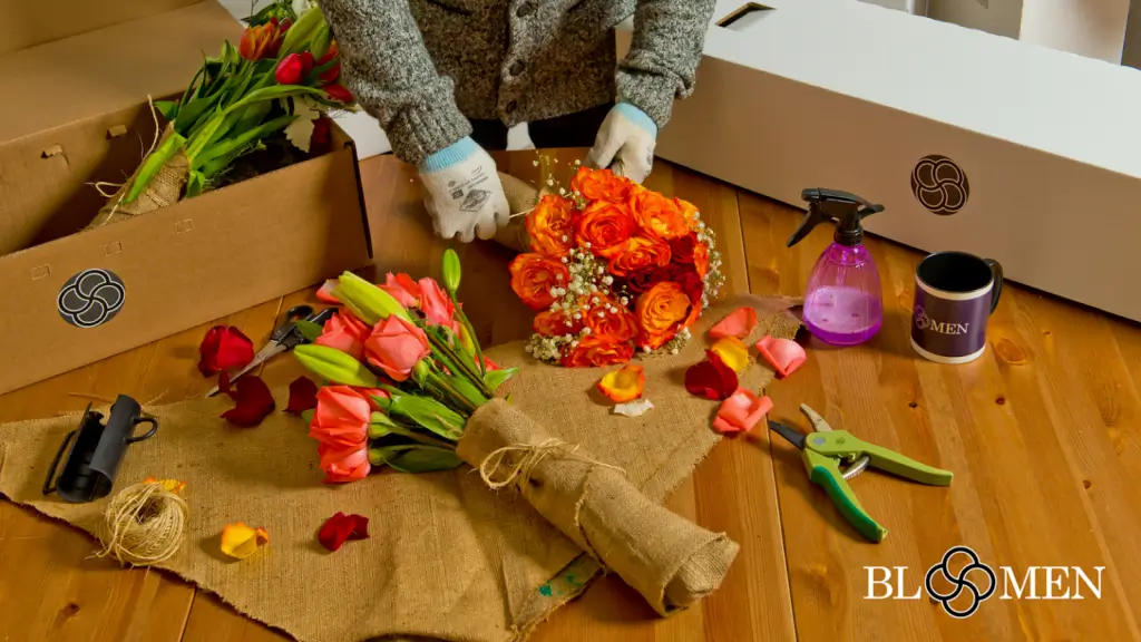 A Bloomen florist in gloves wrapping a bouquet of orange roses, with cut stems, twine, secateurs and branded delivery boxes on the bench