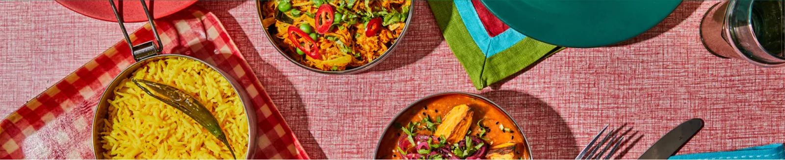 Overhead view of DabbaDrop curries, rice and salad served in stainless steel tiffin tins on a pink cloth