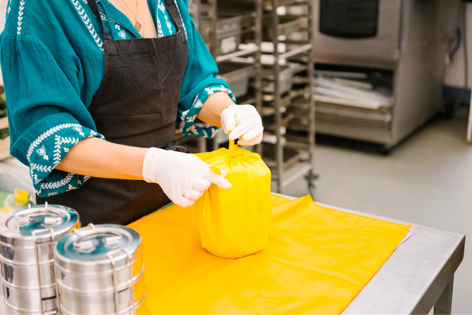 A DabbaDrop kitchen worker in gloves knotting a yellow cloth wrap around a packed tiffin tin