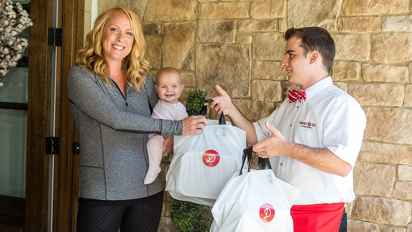 A Donovan's Dish driver handing two bags of meals to a customer holding a baby at her front door