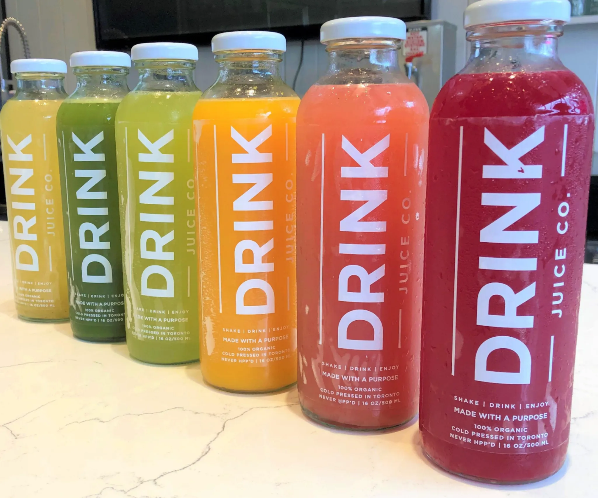 Six Drink Juice Co. bottles lined up on a counter, running from pale yellow through green and orange to deep red