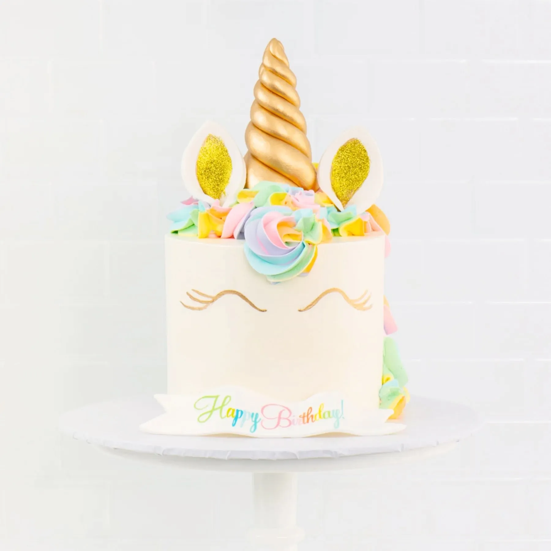 A Sweet E's unicorn birthday cake with a gold fondant horn, pastel buttercream mane and a Happy Birthday! banner