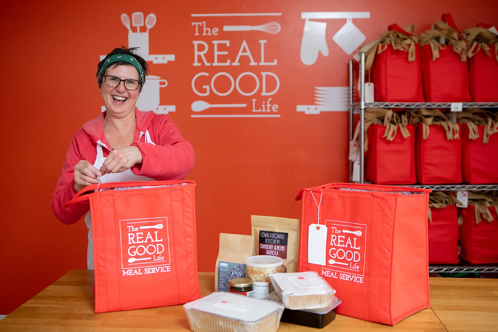 The Real Good Life's owner laughing as she packs an insulated meal tote, with racks of filled red totes behind her